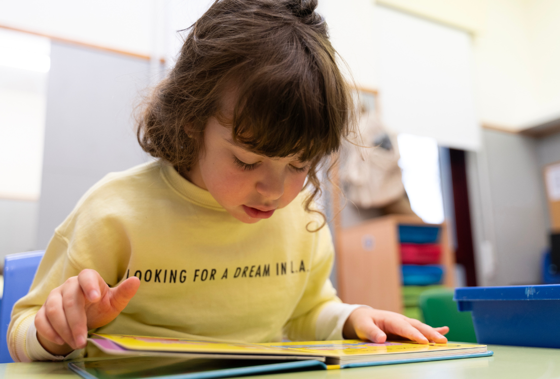 Una niña con una sudadera amarilla lee un libro sentada en una mesa