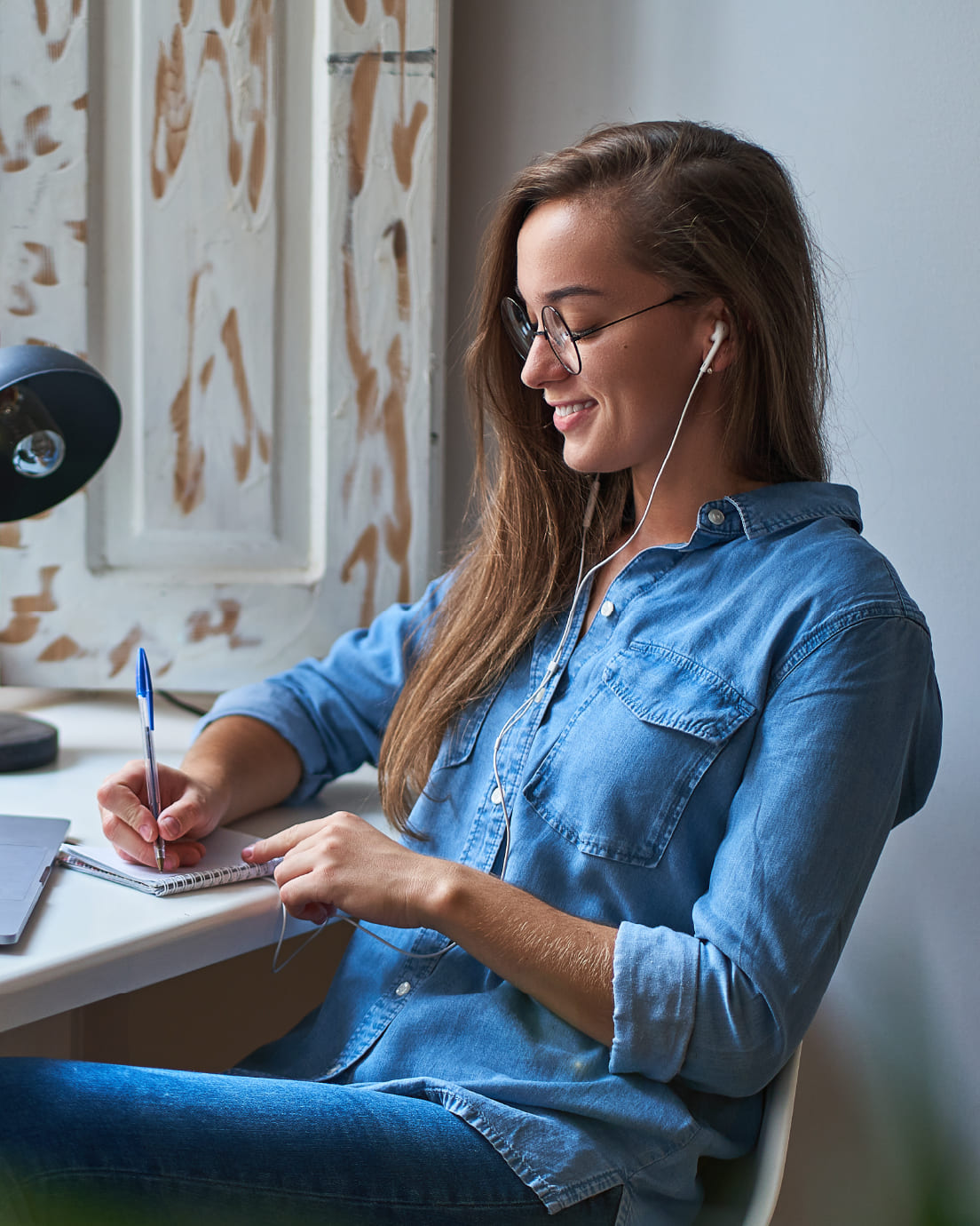 Chica joven sentada en su escritorio escuchando con cascos, escribiendo en una libreta.