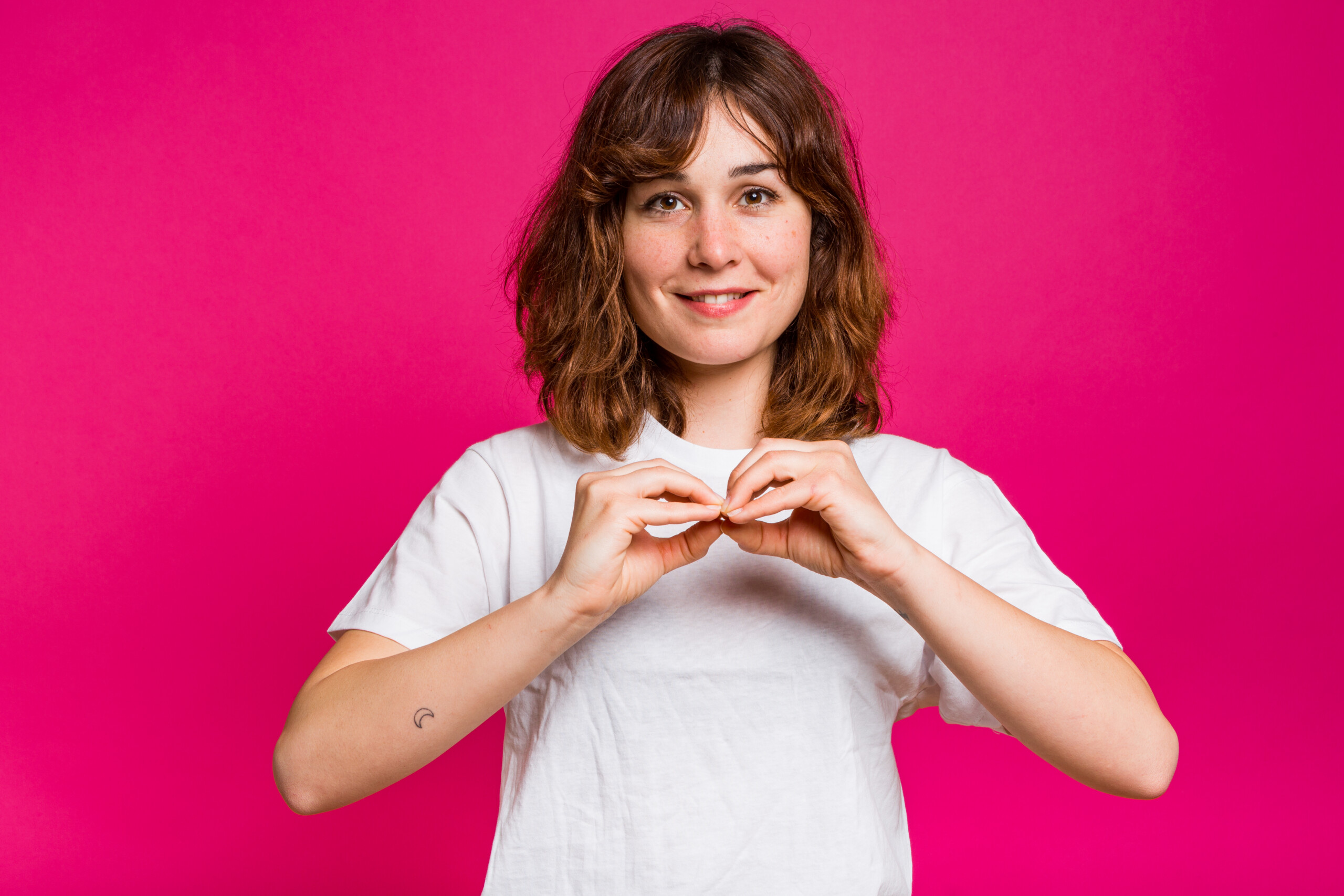La actriz Nadia de Santiago haciendo un corazón con las manos y sonriendo a la cámara sobre un fondo rosa