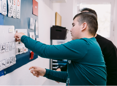 Un chico pegando un papel sobre una pizarra con su persona de apoyo al lado