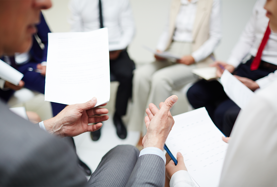 Hombres con traje y corbata sentados en círculo y sujetando unos documentos con las manos