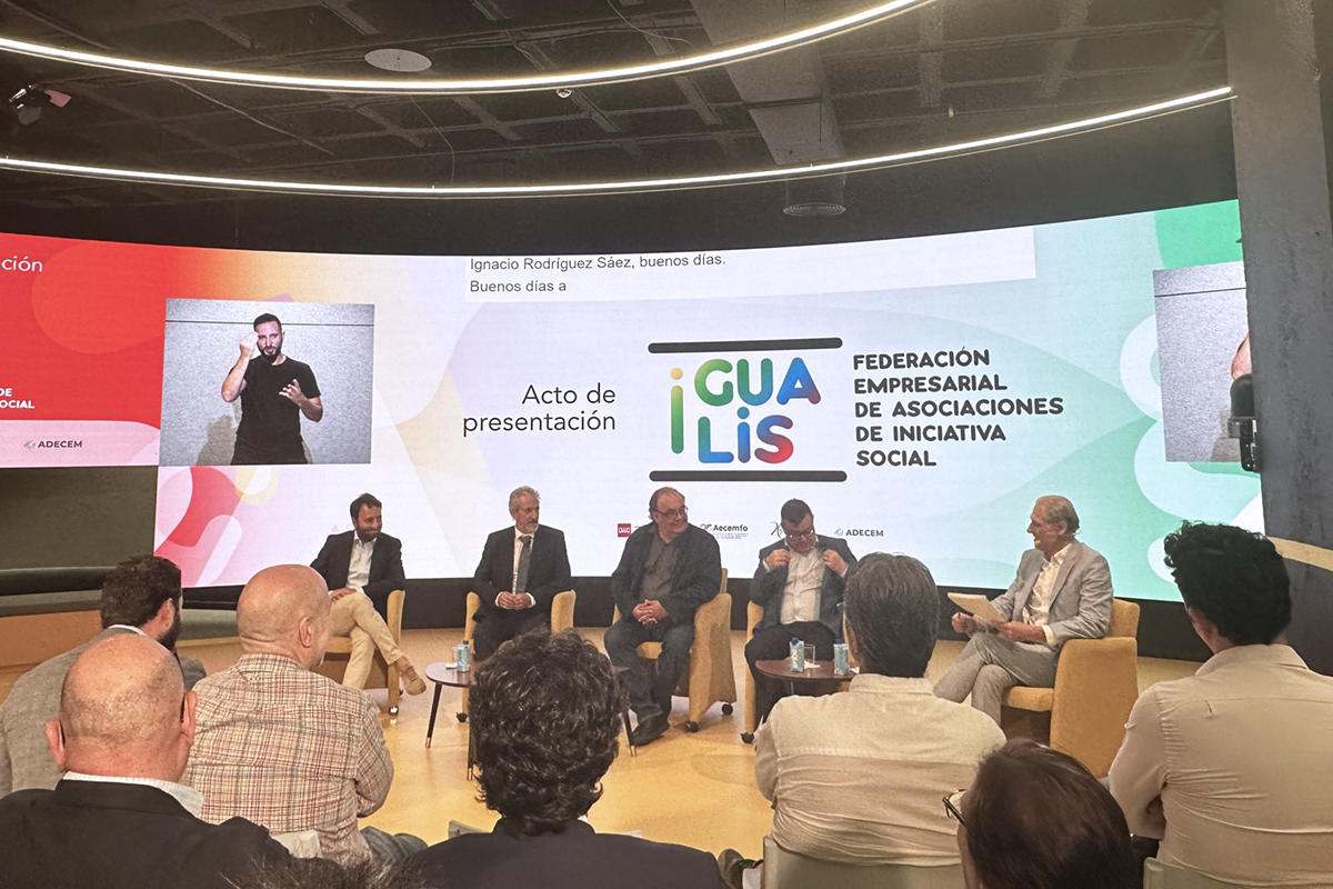 Cuatro personas sentadas en el centro realizando la presentación, con una pantalla grande detrás y un grupo de personas atendiendo