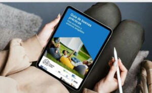 Unas persona sujeta entre sus manos una tableta electrónica con la portada de la guía de buenas prácticas sobre ocio y entornos amigables del proyecto Rumbo. En la foto hay un grupo de personas sobre el césped.