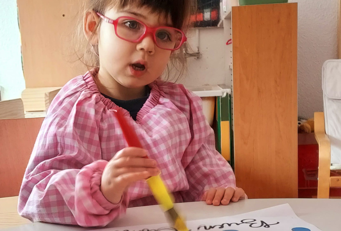 Una niña pintando sentada en un mesa con un babi de cuadros rojos y blancos