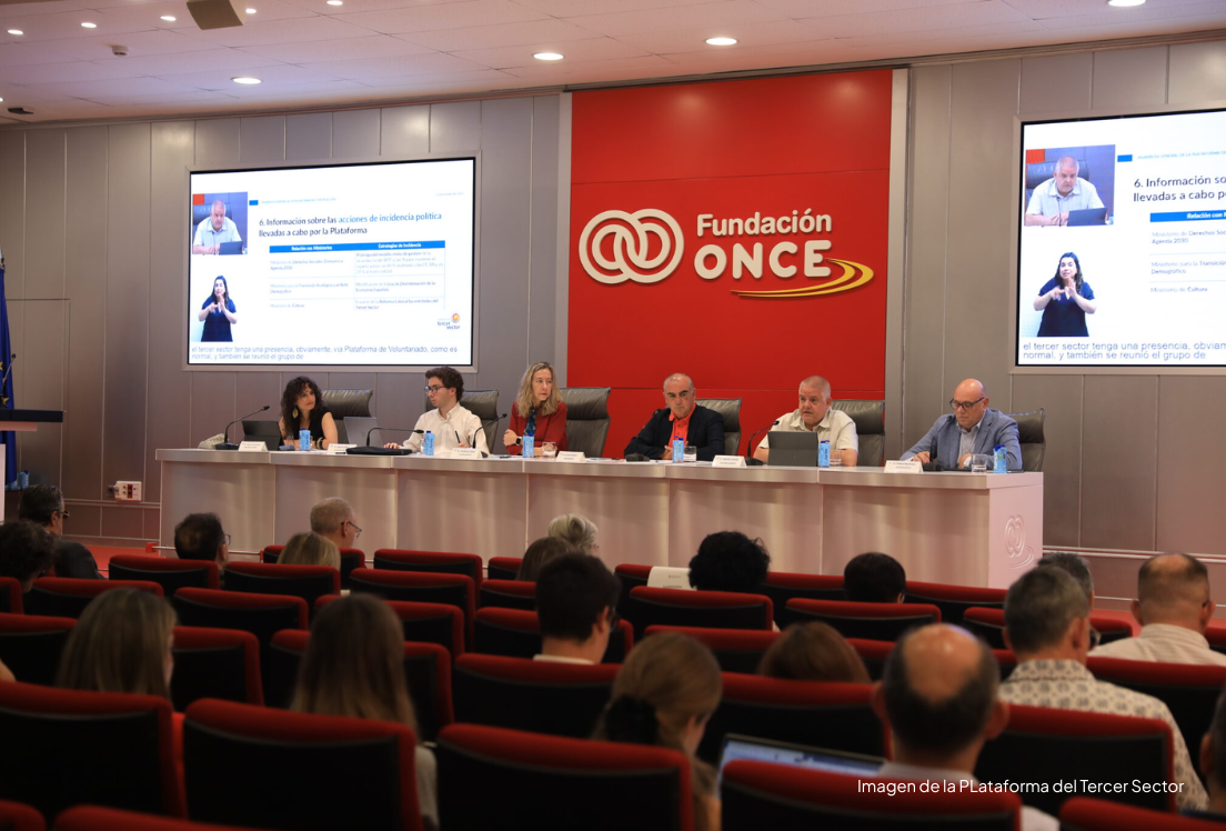 Un salón de actos, con una mesa presidencial con varias personas sentadas y dos pantallas a los lados.