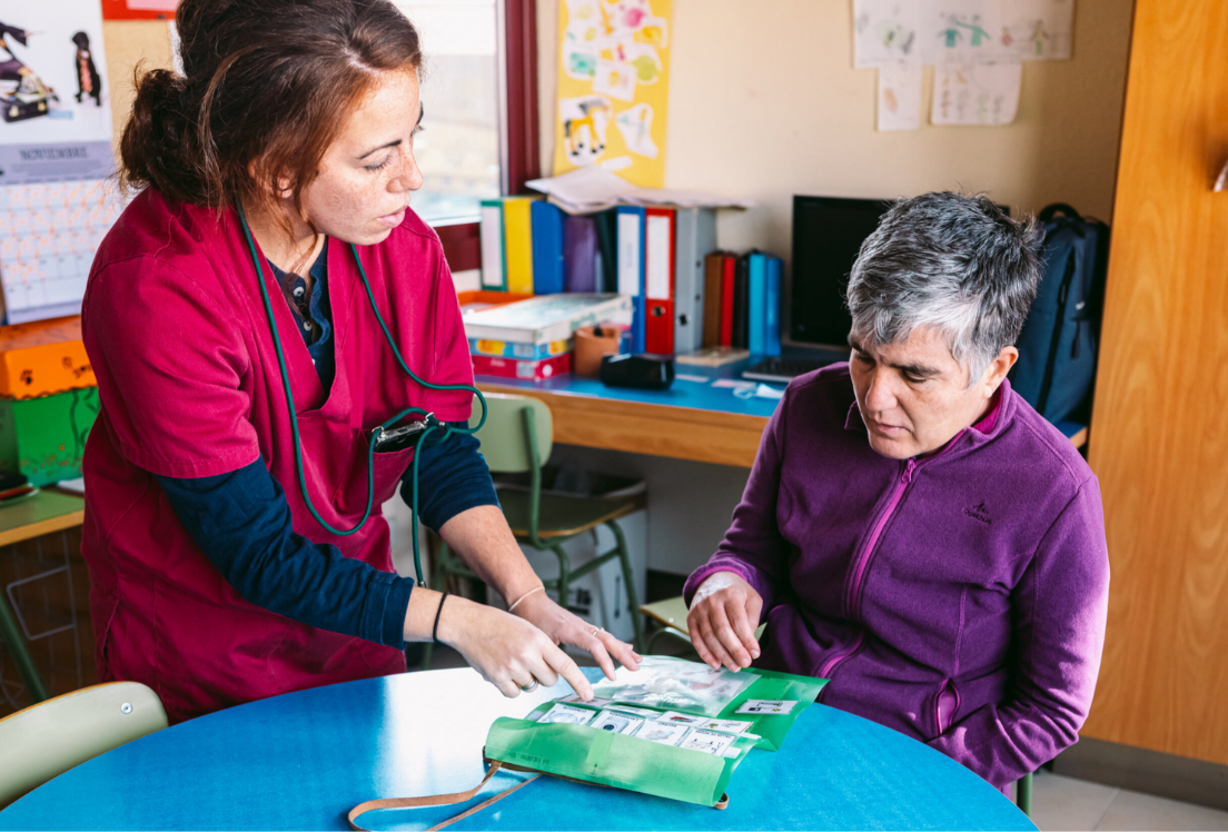 Una persona sentada a una mesa de un aula, señala en un libro de pictogramas con la ayuda de otra persona que está a su lado, de pie