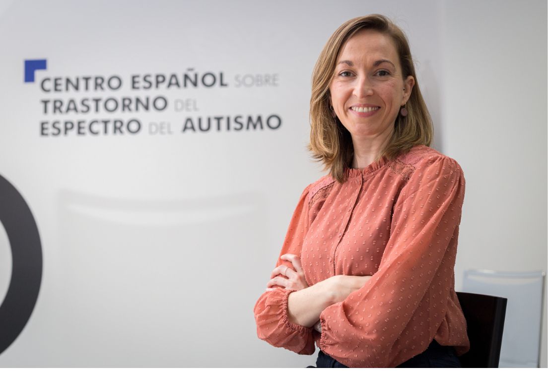 Fotografía de la coordinadora del Centro Español de Autismo, María Luisa Pinos López, de pie con logo del Centro desenfocado al fondo.